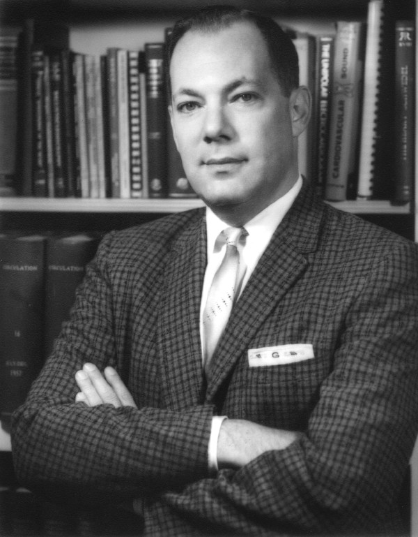 Black-and-white portrait of Marvin Gasster, MD, in a checked jacket with arms crossed, standing in front of shelves of medical books.